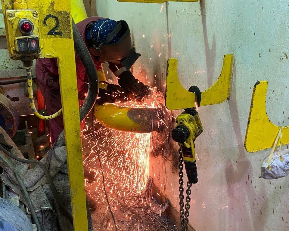 Coded welder grinding a repair inside an offshore vessel compartment, sparks visible, wearing welding shield and protective gloves